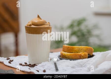 Ein Glas leckeres Dalgona Getränk mit Schlagkaffee und Milch auf dem Tisch im Café zum Frühstück mit Kuchen Stockfoto