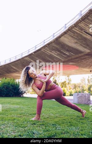 Ganzkörper-Seitenansicht der schlanken, flexiblen Frau im aktiven Ohr Machen rotierten Halbmond Ausfallschritt Pose beim Üben Yoga auf Grün Rasen im Park Stockfoto