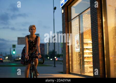 Frau in Kleid reitet Fahrrad in der Nacht Stadt in der Nähe von Geschäft Stockfoto