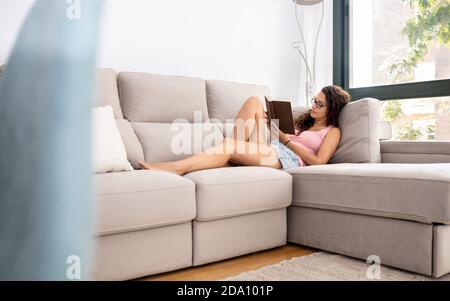 Seitenansicht einer nachdenklichen Frau, die im Wohnzimmer auf der Couch liegt Zimmer und genießen Sie das Lesen interessante Buch Stockfoto