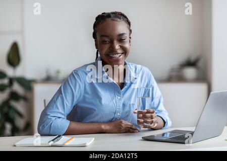 Happy Young African American Frau Hält Glas Wasser, Sitzen Am Arbeitsplatz Stockfoto