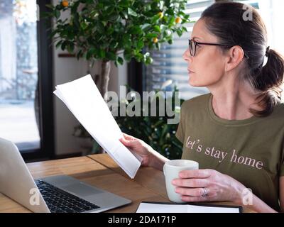 Kaukasische Frau mittleren Alters trägt eine Brille und arbeitet zu Hause mit einem Laptop und Dokumenten. Bleiben Sie zu Hause während covid-19 Quarantäne. Stockfoto