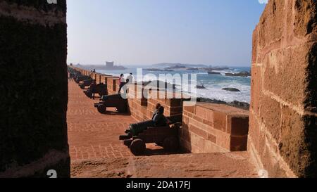 Touristen sitzen auf alten Kanonen und genießen die salzige Meeresbrise des wilden Atlantischen Ozeans auf der historischen Mauer in der Medina von Essaouira, Marokko. Stockfoto