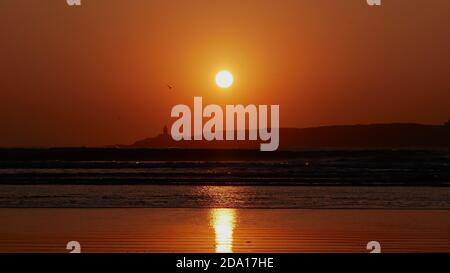 Atemberaubender Sonnenuntergang mit einem orangefarbenen dramatischen Himmel und der hellen Sonne über Mogador Island am Horizont mit brechenden Wellen und gelbem Sonnenlicht. Stockfoto