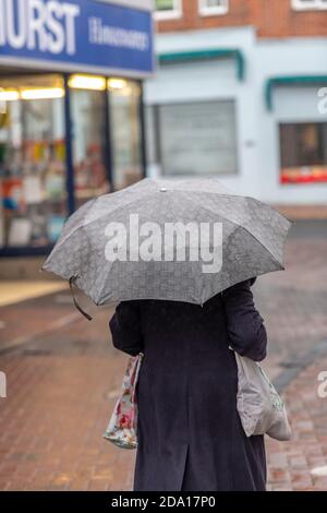 Eine Dame, die einen Wintermantel trägt und einen Regenschirm hält, der an einem regnerischen Herbsttag eine lange Straße im Stadtzentrum führt. Stockfoto