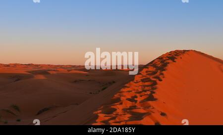 Gipfel einer riesigen orangefarbenen Sanddüne im Abendlicht mit Fußabdrücken im Sand in der Wüste von Erg Chebbi bei Merzouga, Marokko, Afrika. Stockfoto