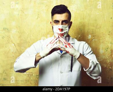 Chirurg mit stilvollen Haarschnitt in Maske mit Blut gefärbt hält seine Hände in Handschuhen zusammen auf Hintergrund beige Farbe Wand Stockfoto