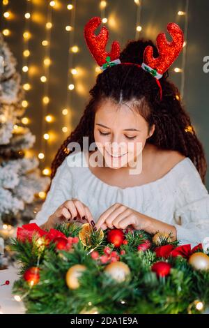 Schön lächelnd afro Haar Frau trägt weiß gestrickt Pullover machen Weihnachtskranz im dekorierten Raum mit Loghts auf Hintergrund Stockfoto