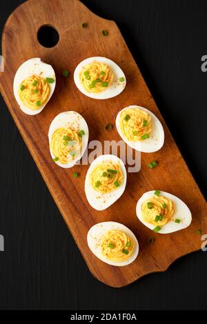 Hausgemachte Deviled Eier mit Schnittlauch auf einem rustikalen Holzbrett auf einer schwarzen Oberfläche, Draufsicht. Flachlage, über Kopf, von oben. Stockfoto