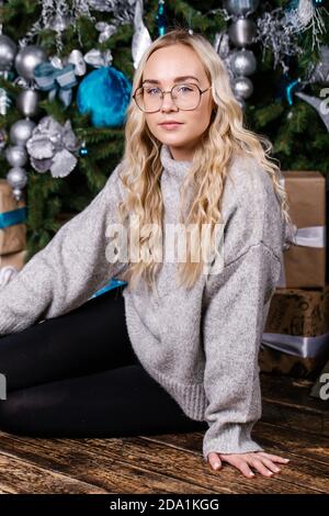 Schönes Mädchen trägt Pullover mit Brille auf dem Hintergrund eines Weihnachtsbaums. Winterferien, Feiern und Menschen Konzept. Stockfoto