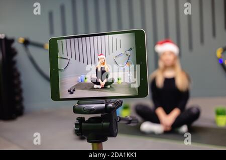 Frau in Santa hat Fitness-Trainer Aufnahme Video-Blog mit Kamera auf Stativ. Live-Streaming, Social Media, Netzwerk, Blogging, Trainingskonzept. Stockfoto