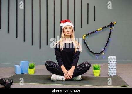 Ein Mädchen in einem Weihnachtsmann Hut tun Fitness-Übungen in der Turnhalle, trägt schwarze Sportuniform, Weihnachten Fitness-Aktivität. Neujahr. Weihnachten, Feiertage, Fi Stockfoto