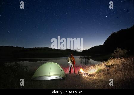 Mann, der in der Nähe von beleuchteten Zelt und Lagerfeuer am See unter Sternenhimmel im Nebrodi Park, Sizilien, steht Stockfoto