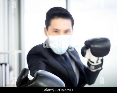 Verschwommen von asiatischen Geschäftsmann im Anzug und schützende Gesichtsmaske und Fokus auf die Hand tragen Boxhandschuhe im Büro. Mann mit Herausforderung trägt Fäustling, Pfosten Stockfoto