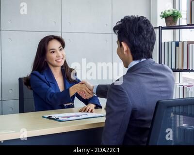 Asiatische Geschäftsfrau und Geschäftsmann tragen Anzug schütteln Hände im Büro. Lächelnde Managerin, die am Schreibtisch im Büro sitzt und mit einem jungen Mann handgeschüttelt Stockfoto