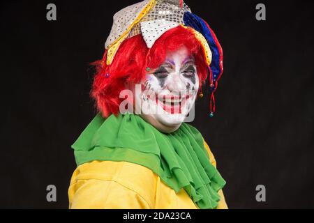 Ein schrecklicher Clown mit einem furchtbaren Make-up und Hut auf dem Kopf lachend auf schwarzem Hintergrund Stockfoto