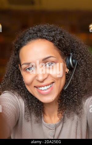 Vertikales Foto einer afroamerikanischen Frau mit einem Headset macht einen Online-Anruf. Sie schaut auf die Kamera und lächelt. Stockfoto