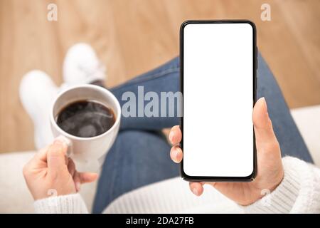 Frauenhand hält weißes Mobiltelefon mit leerem Desktop-Bildschirm und Kaffeetasse. Draufsicht Mockup Bild der Frau hält schwarzes Handy mit leer Stockfoto