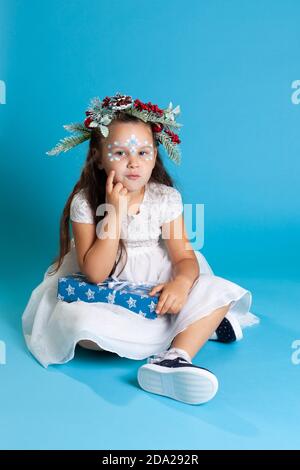 Nachdenkliches Mädchen in einem weißen Kleid, das einen Kranz und Turnschuhe trägt, die auf dem Boden sitzen und einen Finger an ihr Gesicht halten, isoliert auf einem Himmelshintergrund Stockfoto