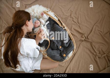 Mama und Baby, die in einer Weidenwiege in einem warmen Strickmütze unter einer warmen Decke mit einem Spielzeug im Griff süß schläft. Gewellter Stoff auf dem Hintergrund Stockfoto