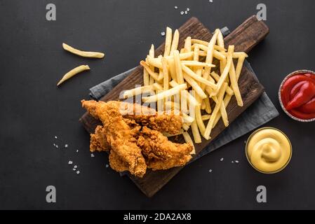 Fisch und Pommes. Frittiertes Fischfilet und mit pommes frites auf schwarzem Tisch mit Saucen serviert. Traditionelles britisches Street Food, Draufsicht, Kopierraum. Stockfoto