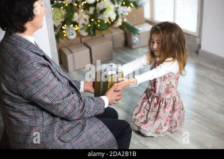 Oma schenkt ihrer Enkelin ein Weihnachtsgeschenk. Das Mädchen streckt ihre Hände auf die Kiste mit dem Bogen. Neujahr, Familienurlaub, communicatio Stockfoto