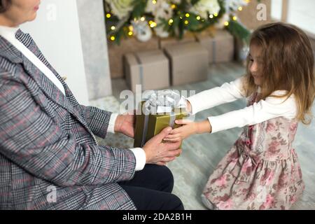 Oma schenkt ihrer Enkelin ein Weihnachtsgeschenk. Das Mädchen streckt ihre Hände auf die Kiste mit dem Bogen. Neujahr, Familienurlaub, communicatio Stockfoto