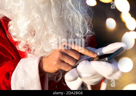 Die Hand des Weihnachtsmannes ohne Fäustling bewegt seinen Finger über den Smartphone-Bildschirm. Langer weißer Bart, roter Anzug. Moderner russischer Großvater Frost. Stockfoto