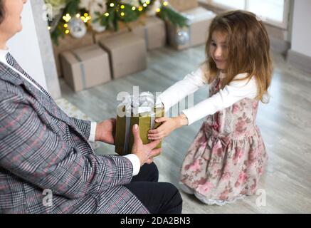 Oma schenkt ihrer Enkelin ein Weihnachtsgeschenk. Das Mädchen streckt ihre Hände auf die Kiste mit dem Bogen. Neujahr, Familienurlaub, communicatio Stockfoto