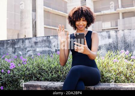 Junge afroamerikanische Frau, die ein Mobiltelefon benutzt und einen Videoanruf macht, während sie auf einer Bank im Park sitzt. Weibliche schwarze Joggerin, die sich ausruhte und die Hände bei der Kamera im Freien winkte Stockfoto