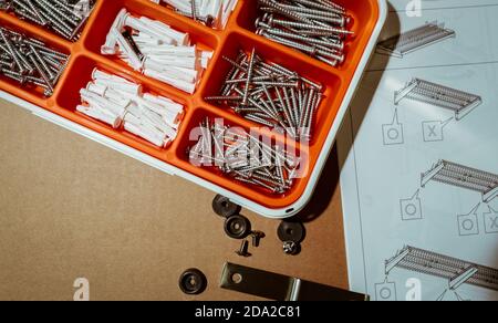 Schrauben und Wandstecker in orangefarbener Kunststoffbox. Werkzeugkasten von oben auf braunem Hintergrund mit Montageanleitung für Spültrockner. Satz Schrauben und Stecker. Stockfoto