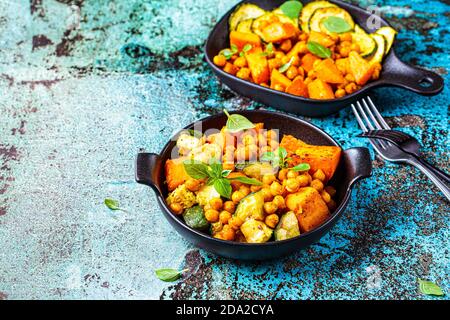 Gebackene Süßkartoffeln, Zucchini und Kichererbsen in gusseisernen Pfannen, blauer Hintergrund. Gebackenes Gemüse. Gesunde Ernährung Konzept. Stockfoto