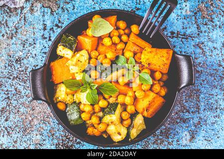 Gebackene Süßkartoffeln, Zucchini und Kichererbsen in gusseisernen Pfannen, blauer Hintergrund. Gebackenes Gemüse. Gesunde Ernährung Konzept. Stockfoto