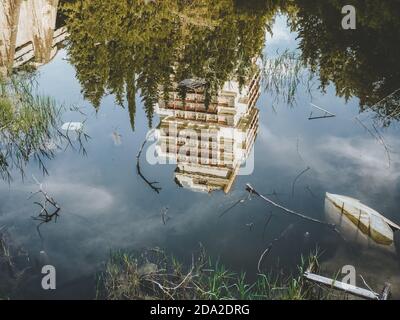 Ein hohes verlassenes Gebäude, umgeben von einem Wald, spiegelt sich wider Im grünen Wasser eines mit Gras bewachsenen Teiches Und übersät mit Müll Stockfoto