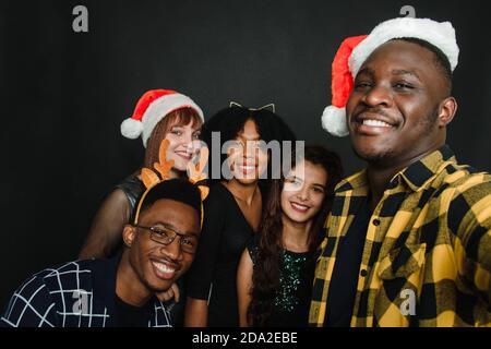 Eine Gruppe von Freunden verschiedener Nationalitäten machen ein Weihnachts-Selfie im Studio. Junge Männer und Frauen in weihnachtsmännern und Hirschgeweihen haben Spaß zusammen. Stockfoto