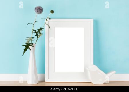 Eleganter vertikaler weißer Bilderrahmen mit mattem Mockup, Schablone mit Kugeldisteln in Vase und stilisiertem Keramikvogel vor pastellblauer Wand. B Stockfoto