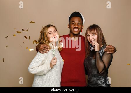 Drei multiethnische Freunde feiern Weihnachten und Neujahr im Studio auf beigem Hintergrund. Zwei kaukasische junge Frauen und ein schwarzer Mann lächeln, während sie in die Kamera schauen. Stockfoto