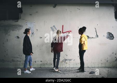 Rückansicht einer Gruppe von Teenagern, die drinnen in einem verlassenen Gebäude stehen, mit Sprühfarbe an der Wand. Stockfoto