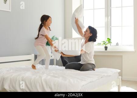 Fröhliche Mama und nette kleine Tochter kämpfen mit Kissen auf Breites Bett zu Hause Stockfoto