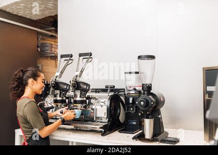 Weibliche Barista, die bei der Kaffeemaschine steht, während sie im Café arbeitet. Frau in der Schürze, die Kaffee zubereitet. Stockfoto
