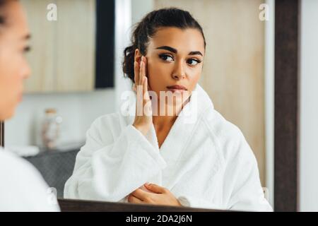Mixed-Race-Frau im Badezimmer stehend berühren ihr Gesicht Beim Blick auf einen Spiegel Stockfoto