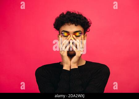 Homosexuell Mann trägt Regenbogen farbigen Lidschatten. Androgynen Mann hält sein Gesicht mit geschlossenen Augen auf rotem Hintergrund. Stockfoto