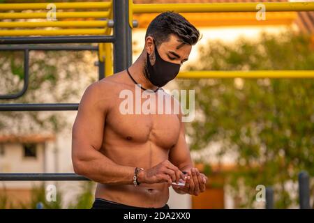 Junger Latino Mann ohne Hemd und schwarze Hose und Maske, die seine Hände während des Trainings mit hydroalkoholischem Gel desinfiziert Ein Calisthenics Park Stockfoto