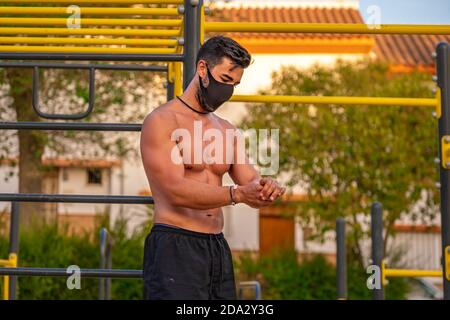 Junger Latino Mann ohne Hemd und schwarze Hose und Maske, die seine Hände während des Trainings mit hydroalkoholischem Gel desinfiziert Ein Calisthenics Park Stockfoto