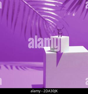 Eine kleine Figur eines goldenen Hirsches - ein Symbol des neuen Jahres auf zwei weißen Quadraten, im Stil des Monumentalismus, auf einem gelben Hintergrund, hinter den Blättern einer Palme. Stockfoto