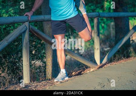 Junger Kaukasianer mit Maske und blauem Hemd beim Dehnen Auf dem Zaun eines Parks mit Bäumen in der Hintergrund Stockfoto