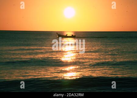 Sonnenuntergang über dem Meer mit einem kleinen Fischerboot und zwei Fischern über dem Wasser und vor der Sonnenlinie; Sonnenlicht innerhalb der Wolken Stockfoto