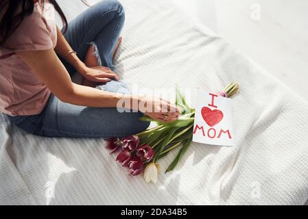 Draufsicht auf die Frau, die auf dem Bett mit Blumenstrauß sitzt Von Blumen und Postkarte mit Herzbildern darauf Stockfoto