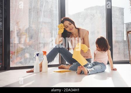 Müde Frau mit ihrer Tochter sitzt auf Fensterbank mit Flaschen mit Reinigungsspray und mit Handschuhen Stockfoto