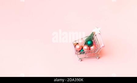 Einige festliche Bälle und Miniatur-Weihnachtsbaum in Spielzeug Supermarkt Trolley auf Papier rosa Hintergrund. Neujahrs- und Weihnachtseinkaufskonzept. Stockfoto